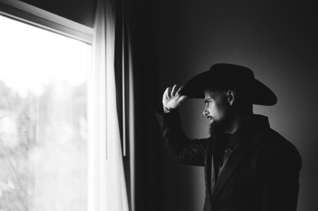 35mm black and white film photograph of a groom in a black cowboy hat standing at a window and tipping the brim, lit only by the daylight beside him. Shot on CineStill BwXX before a wedding in Easton, Pennsylvania, by film wedding photographer Drew Ferguson.