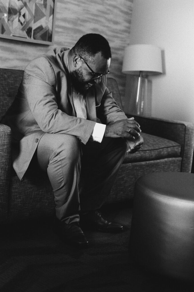 35mm black and white film photograph of a groom in a light suit sitting on a hotel couch, leaning forward with his hands clasped and laughing at something off camera. Shot on CineStill BwXX before Stephanie and Kevin's wedding at Northampton Valley Country Club, by film wedding photographer Drew Ferguson.