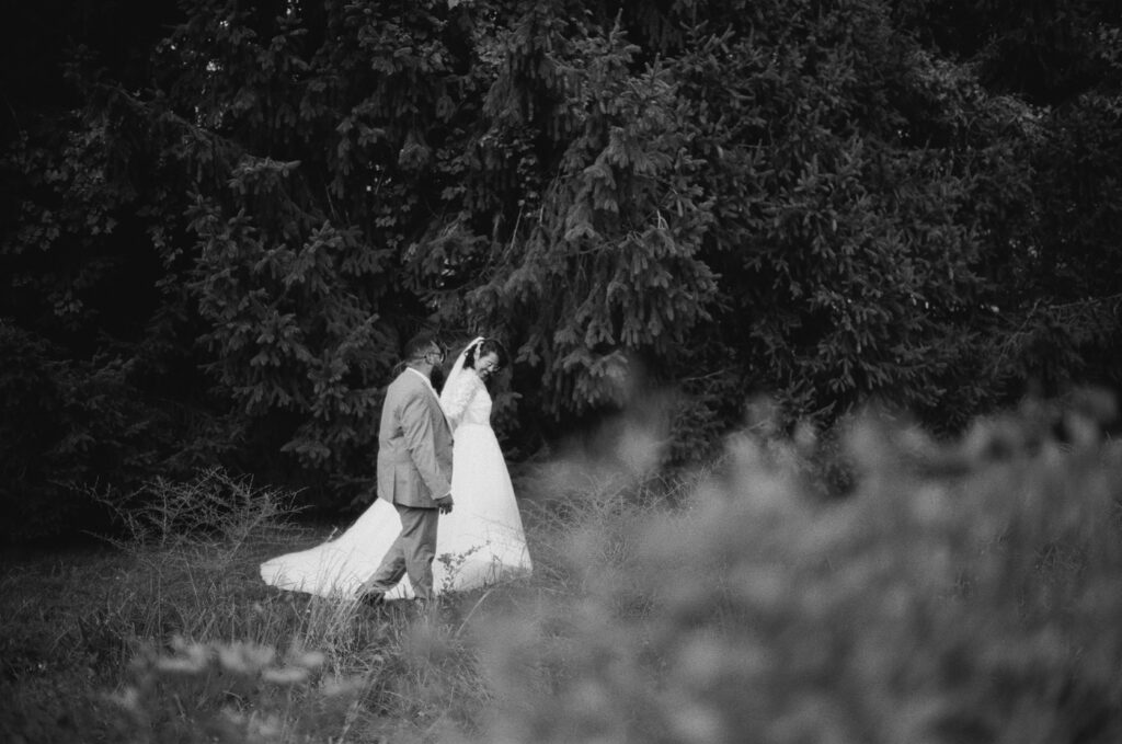 35mm black and white film photograph of a bride and groom walking hand in hand through tall grass in front of a wall of spruce trees, seen through blurred grass in the foreground. Stephanie and Kevin's first look in Warminster, Pennsylvania, shot on CineStill BwXX by film wedding photographer Drew Ferguson.