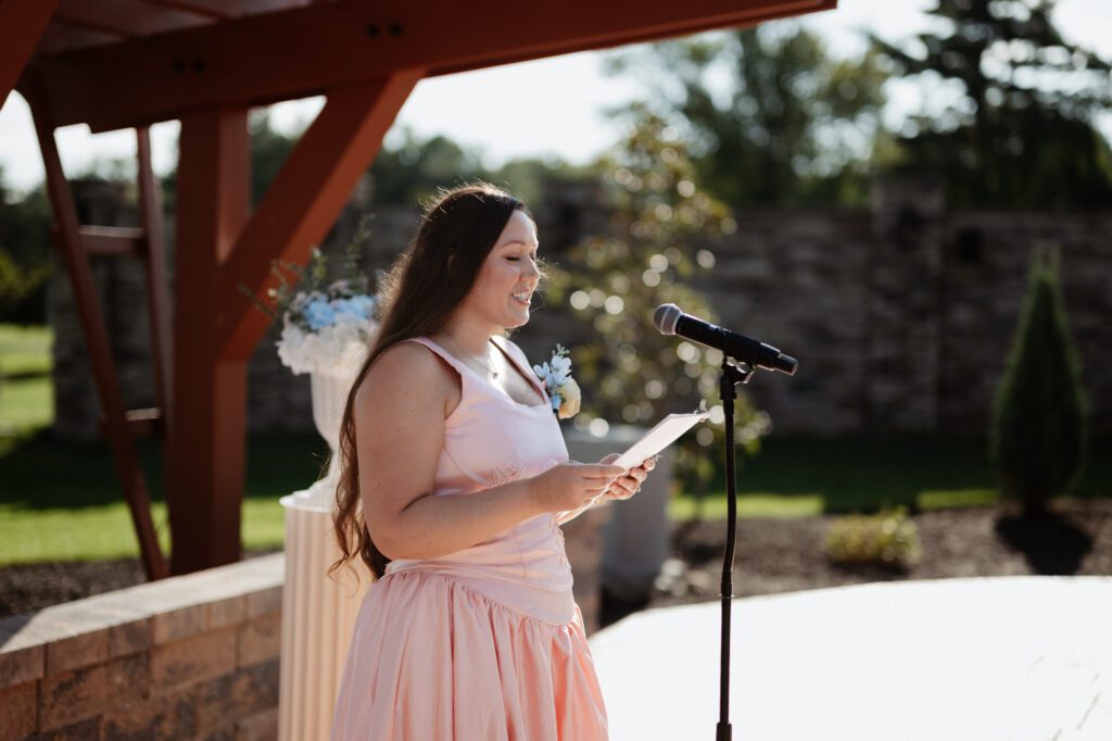 A woman in a blush dress reading from a card at a microphone beneath the red pergola, late afternoon sun behind her. Erin officiating Stephanie and Kevin's wedding ceremony at Northampton Valley Country Club in Richboro, Pennsylvania, by wedding photographer Drew Ferguson.