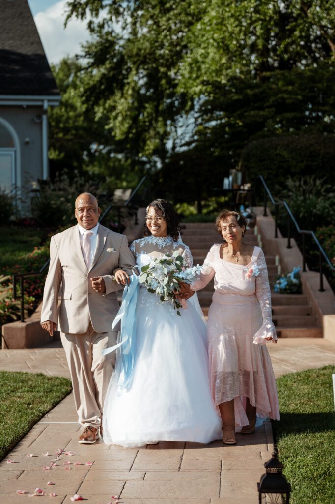 A smiling bride walking down a stone path on the arms of her parents, holding a bouquet of white and blue flowers tied with a long blue ribbon, rose petals scattered ahead of her. The processional at Stephanie and Kevin's wedding at Northampton Valley Country Club in Richboro, Pennsylvania, by wedding photographer Drew Ferguson.