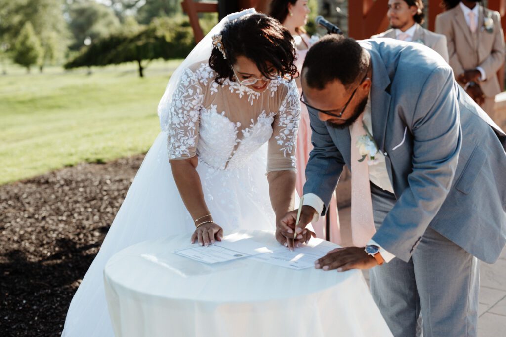 A bride and groom leaning over a small draped table to sign their marriage certificate during the ceremony, the officiant and wedding party behind them under the pergola. Stephanie and Kevin signing their self-uniting marriage license at Northampton Valley Country Club in Richboro, Pennsylvania, by wedding photographer Drew Ferguson.