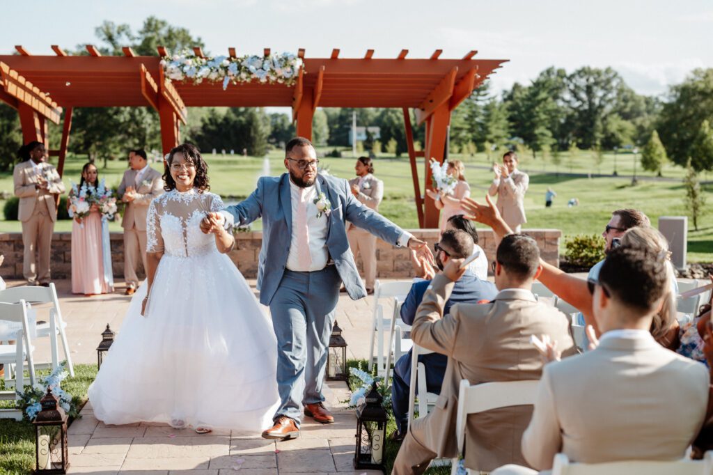 A bride and groom walking back up the aisle after the ceremony, the groom reaching out to slap hands with a guest, the wedding party applauding under the red pergola behind them. The recessional at Stephanie and Kevin's wedding at Northampton Valley Country Club in Richboro, Pennsylvania, by wedding photographer Drew Ferguson.