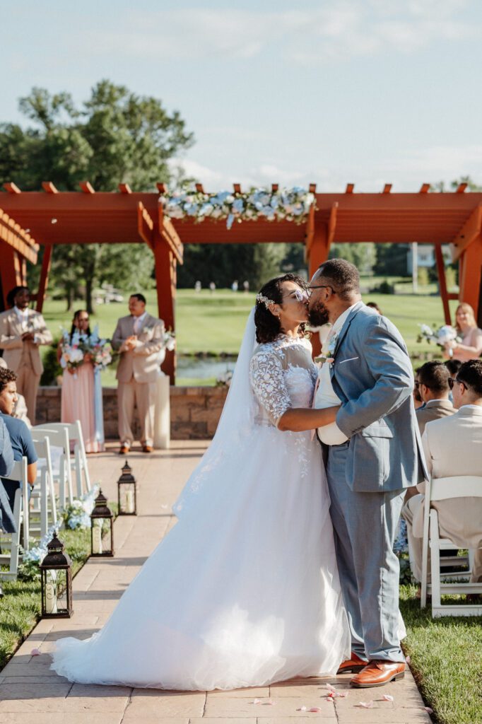 A bride and groom kissing in the aisle in front of the red pergola, the wedding party and seated guests behind them and rose petals on the stone path. Stephanie and Kevin's ceremony at Northampton Valley Country Club in Richboro, Pennsylvania, by wedding photographer Drew Ferguson.