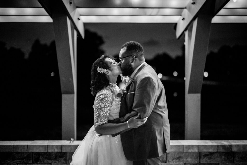Black and white photograph of a bride and groom kissing beneath the lit pavilion at dusk, string lights blurred behind them and the stone wall below. Stephanie and Kevin at the end of the night at Northampton Valley Country Club in Richboro, Pennsylvania, by wedding photographer Drew Ferguson.