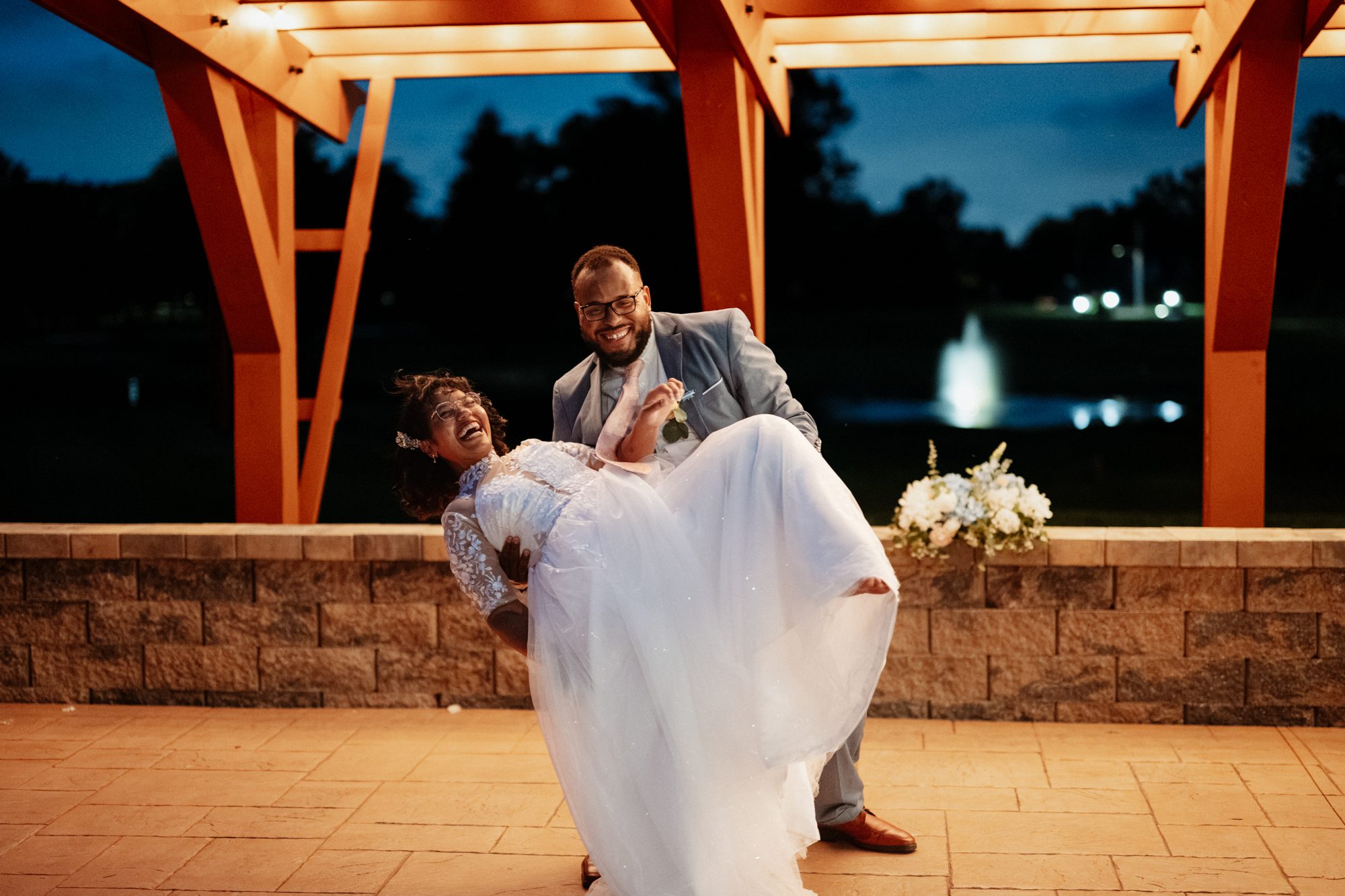 A groom in a light blue suit dipping his laughing bride beneath the lit timber pavilion at blue hour, with the pond fountain glowing behind them. Stephanie and Kevin's wedding at Northampton Valley Country Club in Richboro, Pennsylvania, by wedding photographer Drew Ferguson.