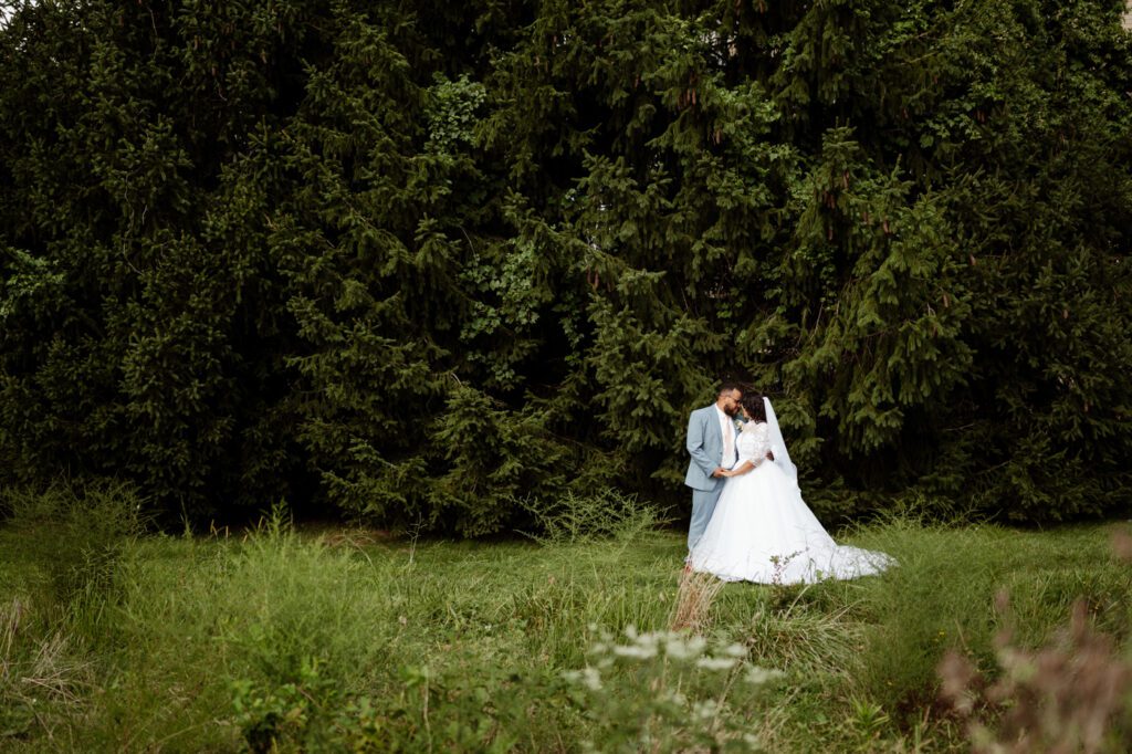 A bride and groom standing close together in a field of tall grass, small against a dense wall of spruce trees, the bride's veil catching the light. Stephanie and Kevin's first look in Warminster, Pennsylvania, before their wedding at Northampton Valley Country Club, by wedding photographer Drew Ferguson.