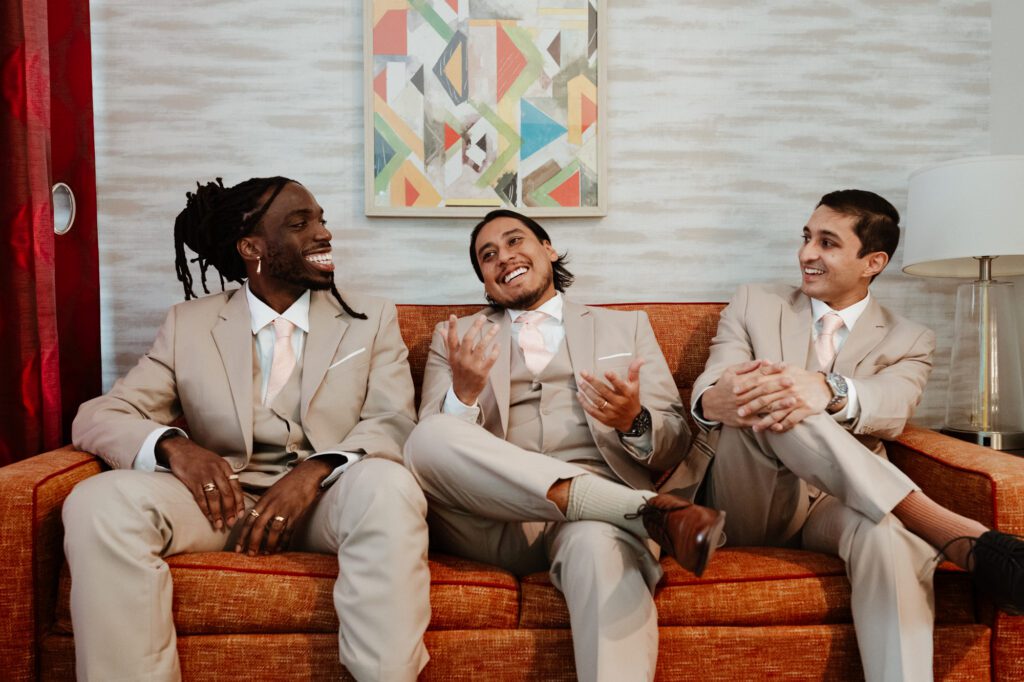 Three groomsmen in tan suits and blush ties laughing together on an orange couch in a hotel suite before the wedding, a geometric print on the wall behind them. Getting ready at the Home2 Suites in Warminster, Pennsylvania, for Stephanie and Kevin's wedding at Northampton Valley Country Club, by wedding photographer Drew Ferguson.