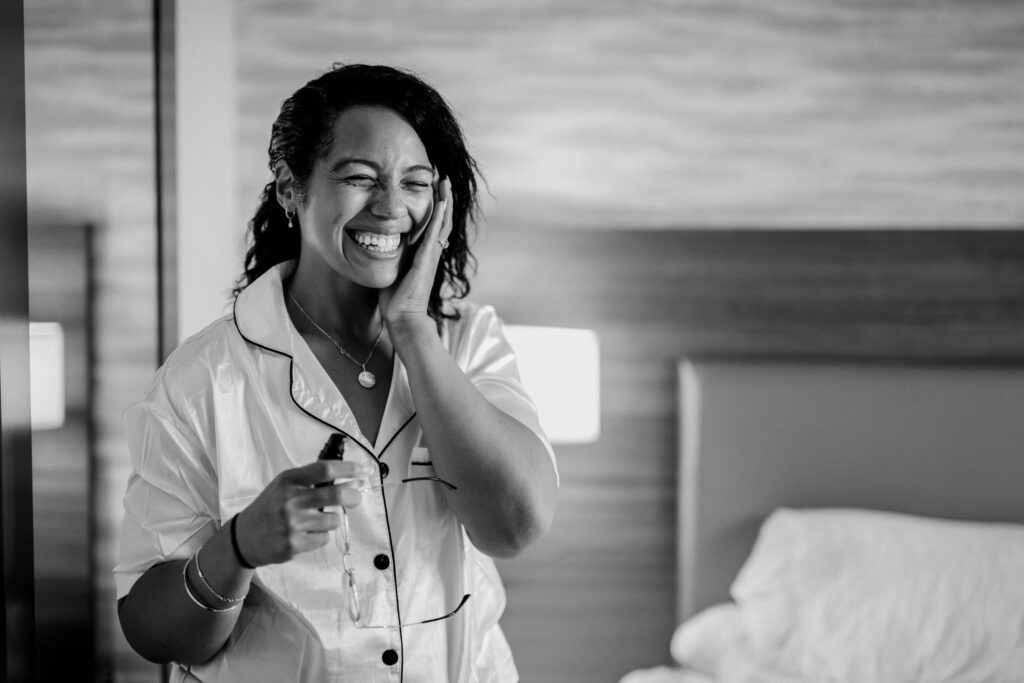 Black and white photograph of a bride in satin pajamas laughing with one hand on her cheek and her glasses in the other, in a hotel room on the morning of her wedding. Stephanie getting ready in Warminster, Pennsylvania, before her wedding at Northampton Valley Country Club, by wedding photographer Drew Ferguson.