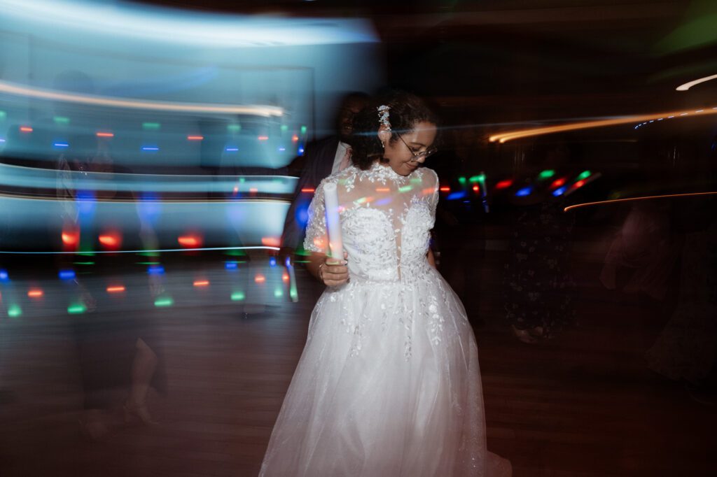 A bride dancing with her eyes down and a foam light stick in hand, streaks of red, green, and blue dance-floor lights blurred across the frame behind her. Stephanie on the dance floor at Northampton Valley Country Club in Richboro, Pennsylvania, by wedding photographer Drew Ferguson.