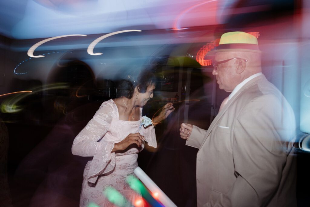 The mother of the bride in a blush lace dress dancing with a man in a tan suit and a neon fedora, dance-floor lights blurred in arcs around them. The reception at Stephanie and Kevin's wedding at Northampton Valley Country Club in Richboro, Pennsylvania, by wedding photographer Drew Ferguson.