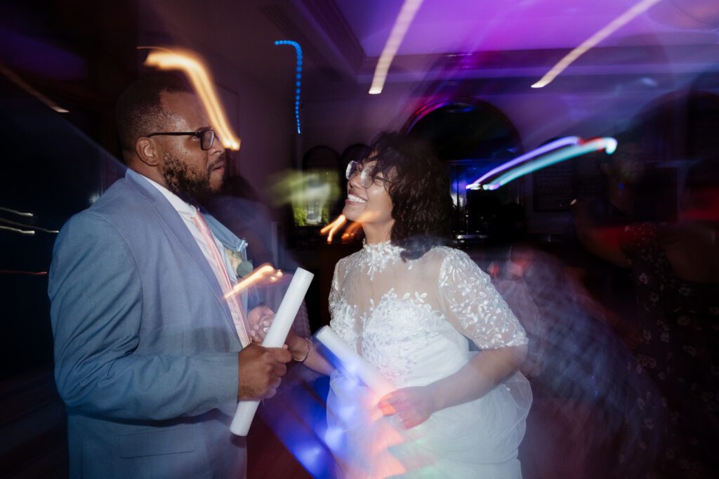 A bride and groom face to face on the dance floor holding foam light sticks, colored light trails streaking across the frame from a slow shutter. Stephanie and Kevin's reception at Northampton Valley Country Club in Richboro, Pennsylvania, by wedding photographer Drew Ferguson.