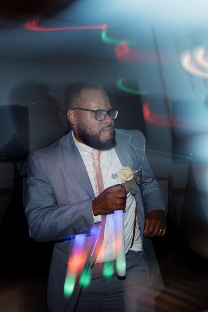 A groom in a light blue suit and blush tie holding a foam light stick on the dance floor, the stick's glow trailing in green, blue, and orange from a slow shutter. Kevin at his reception at Northampton Valley Country Club in Richboro, Pennsylvania, by wedding photographer Drew Ferguson.