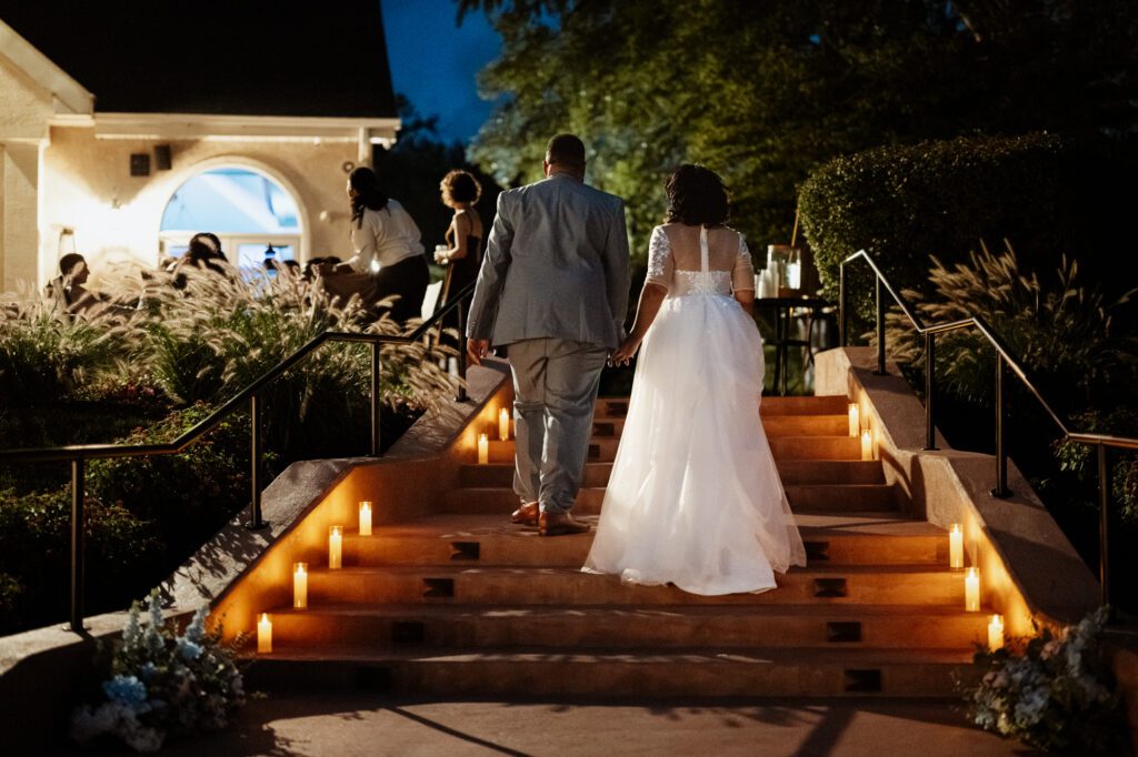 A bride and groom seen from behind, holding hands and climbing stone steps lined with pillar candles toward the reception at dusk, guests and tall grasses ahead of them. Stephanie and Kevin at Northampton Valley Country Club in Richboro, Pennsylvania, by wedding photographer Drew Ferguson.