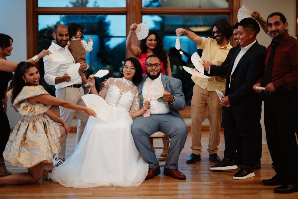 A bride and groom seated in the middle of a table of guests who surround them waving paper fans, one friend kneeling in front with a fan and a small child held on a hip at the back. The song rush at Stephanie and Kevin's reception at Northampton Valley Country Club in Richboro, Pennsylvania, by wedding photographer Drew Ferguson.
