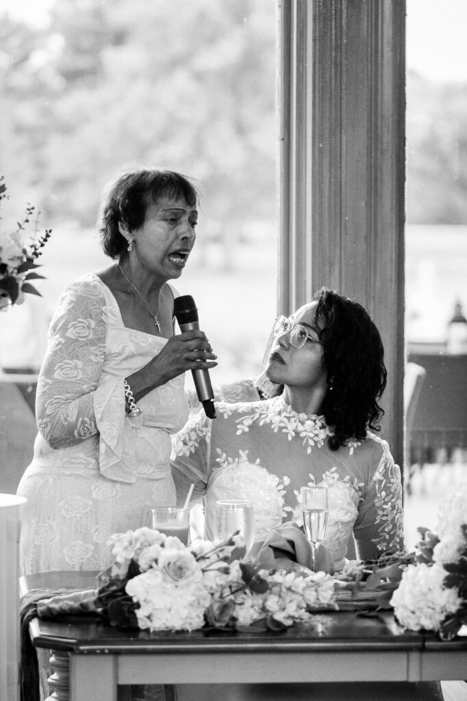 Black and white photograph of the mother of the bride speaking into a microphone beside the sweetheart table while the bride looks up at her, a hand resting on her shoulder. The toasts at Stephanie and Kevin's reception at Northampton Valley Country Club in Richboro, Pennsylvania, by wedding photographer Drew Ferguson.